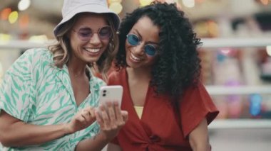 Two laughing multinational young women wearing sunglasses typing on mobile in adventure park