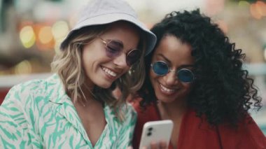 Two smiling multinational young women wearing sunglasses texting on mobile in adventure park