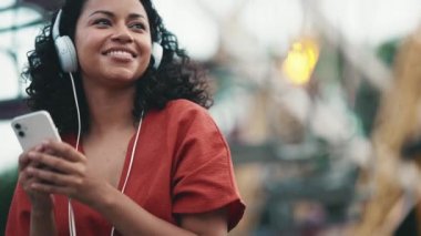 Happy hispanic curly haired woman listening music in headphones while sitting in adventure park