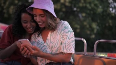Two funny multinational young women typing by phone while riding a ferris wheel in adventure park