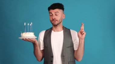 Cheerful brunet man wearing a vest and t-shirt makes a wish while holding a birthday cake in the blue studio