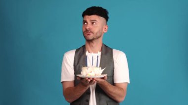 Upset brunet man wearing a vest and t-shirt looking around and at the birthday cake in the blue studio
