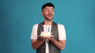 Positive brunet man wearing a vest and t-shirt looking around and blowing out the candles on the birthday cake in the blue studio