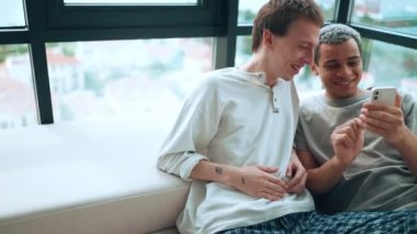 Happy gay couple looking at phone and talking while sitting on the sofa at home