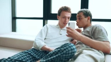 Positive gay couple looking at phone and talking while sitting on the sofa at home
