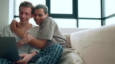 Happy gay couple looking at laptop and talking while sitting on the couch at home