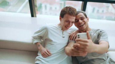 Cheerful gay couple making selfie photo on phone and talking while sitting on the sofa at home