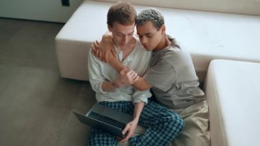 Pretty gay couple looking at laptop and talking while sitting on the floor at home