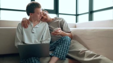 Positive gay couple working on laptop and talking while looking at phone at home