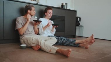 Smiling gay couple eating breakfast and talking while sitting on the floor in the kitchen at home