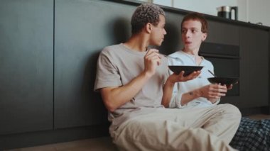Positive gay couple eating breakfast and talking while sitting on the floor in the kitchen at home