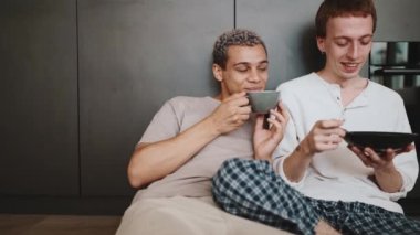Pretty gay couple eating breakfast and drinking coffee while talking on the floor in the kitchen