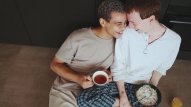 Cheerful gay couple eating breakfast and drinking coffee while talking on the floor in the kitchen