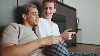 Laughing gay couple eating breakfast and drinking coffee while talking on the floor in the kitchen