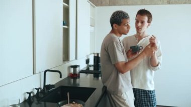 Confident multinational gay couple talking and holding hands while standing in the kitchen