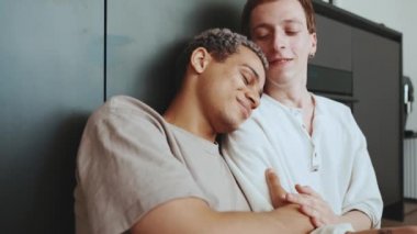 Positive multinational gay couple hugging and kissing while sitting on the kitchen floor
