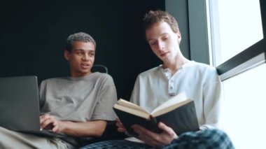 Happy multinational gay couple working on laptop and reading book while talking at home