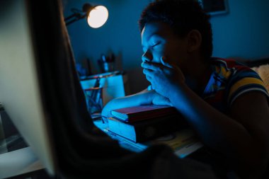 Black boy yawning while doing homework by computer monitor at home