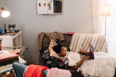 Black boy working with laptop while lying on sofa at home