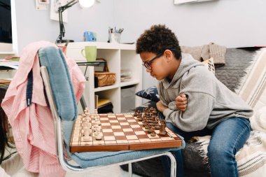 Black boy in eyeglasses playing chess while sitting on sofa at home