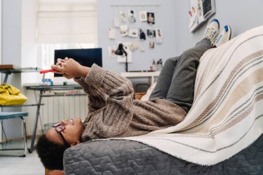 Black boy in eyeglasses laughing and using cellphone while resting at home