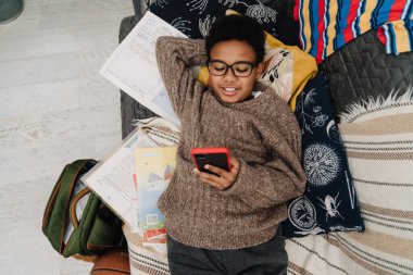 Black boy in eyeglasses laughing and using cellphone while resting at home
