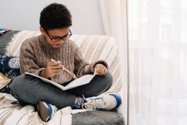 Black boy smiling and doing homework while sitting on sofa at home
