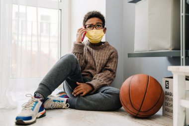 Black boy in face mask talking on cellphone while sitting on floor at home