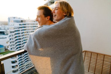 European gay couple wrapped blanket laughing and hugging on balcony at home
