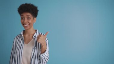 Cheerful hispanic curly haired woman pointing to the copyspace and looking at the camera in the blue studio