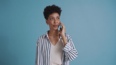 Positive hispanic curly haired woman talking on phone while standing in the blue studio