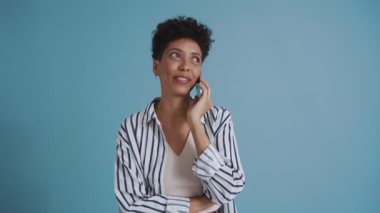 Pleased hispanic curly haired woman talking on phone while standing in the blue studio