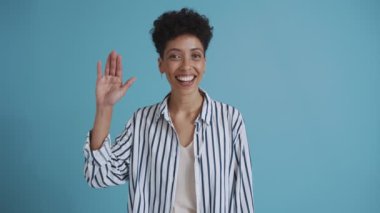 Smiling hispanic curly haired woman showing goodbye gesture at the camera while standing in the blue studio