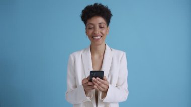 Smiling hispanic curly haired woman wearing white jacket texting by phone in the blue studio
