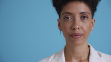 Face of serious hispanic curly haired woman wearing white jacket looking at the camera in the blue studio