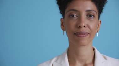 Face of smiling hispanic curly haired woman wearing white jacket looking at the camera in the blue studio