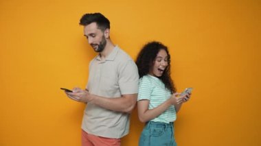 Smiling brunette couple typing on phones while standing back to back in the yellow studio