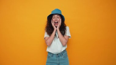Surprised brunette curly haired woman wearing panama hat rejoicing at the camera in the yellow studio