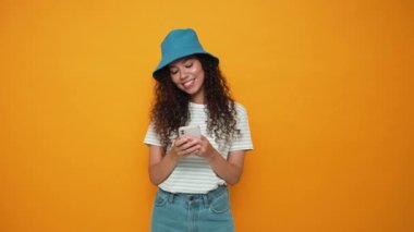 Pretty brunette curly haired woman wearing panama hat typing on mobile in the yellow studio