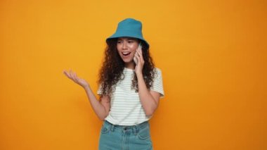 Positive brunette curly haired woman wearing panama hat talking on mobile in the yellow studio