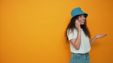 Laughing brunette curly haired woman wearing panama hat talking on mobile in the yellow studio
