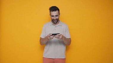 Rejoicing brunet man wearing t-shirt playing video game on mobile while standing in the yellow studio