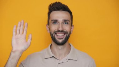 Pretty brunet man wearing t-shirt waving hand at the camera while standing in the yellow studio