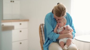 Pleased blonde mother hugging her small child in her arms while sitting in the kitchen