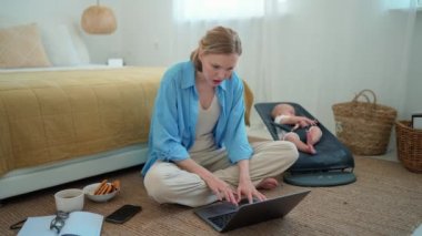 Concentrated blonde mother working on laptop and talking by phone while her small child lying near her indoors at home