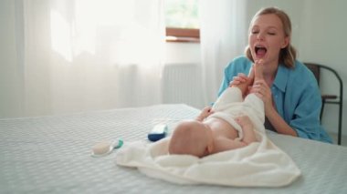 Funny blonde mother talking and playing with legs of her little baby on the bed at home