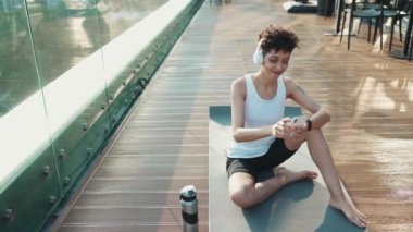 Positive young African sporty woman in headphones typing by phone while sitting on the yoga mat outdoors