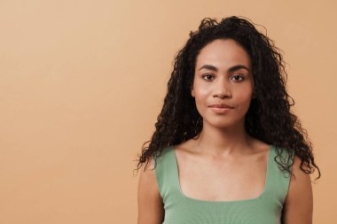 Young black woman with wavy hair posing and looking at camera isolated over beige background