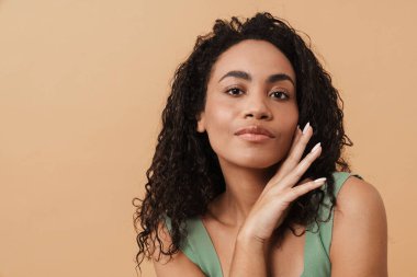Young black woman touching her face and looking at camera isolated over beige background