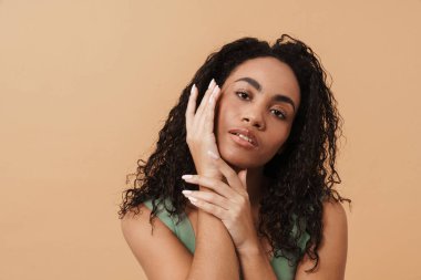 Young black woman touching her face and looking at camera isolated over beige background